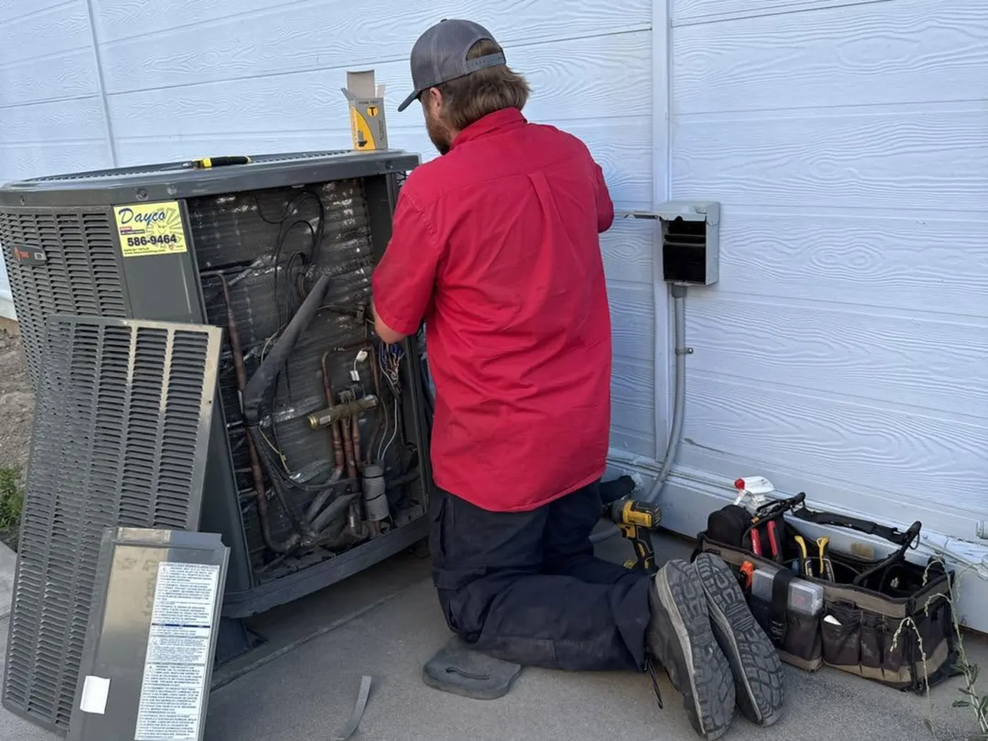 A Dayco technician in a red company shirt kneeling at an outdoor unit with its service panels off, tool bag beside him